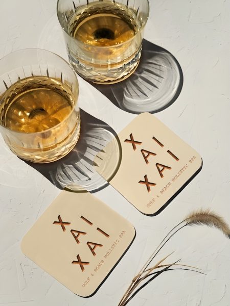 Elegant glass cups with golden drink, name cards and feather on white surface - Xai-Xai hotel